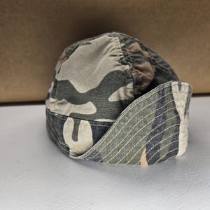 May include: A camouflage print cap in shades of green, brown, and tan. The cap has a curved brim and a soft, unstructured crown. The brim's underside features a striped pattern. The cap is made of a durable fabric.