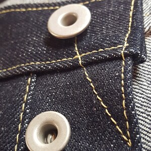 May include: Close-up of dark blue denim fabric with two silver metal rivets. The denim has yellow stitching around the pocket and rivets. The fabric is textured and detailed.