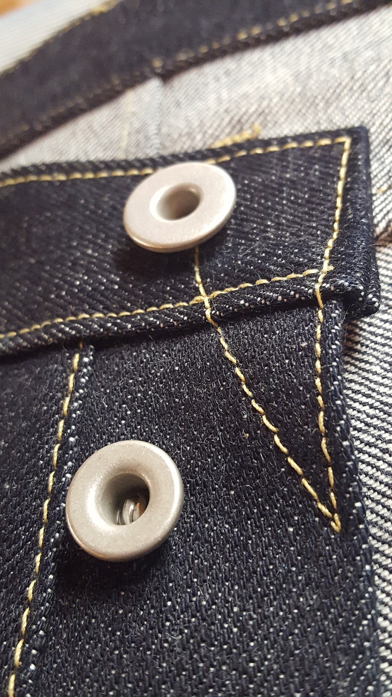 May include: Close-up of dark blue denim fabric with two silver metal rivets. The denim has yellow stitching around the pocket and rivets. The fabric is textured and detailed.