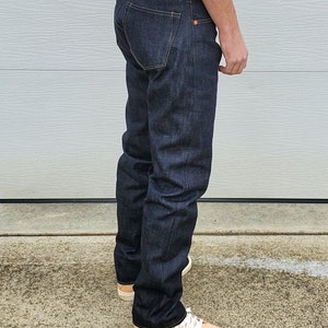 May include: Dark wash denim jeans with a classic five-pocket design. The jeans have a straight leg fit and are worn with light-colored sneakers. The denim appears to be a deep indigo color, with subtle fading.
