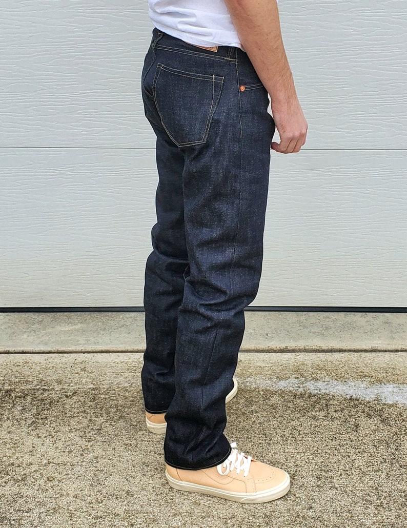 May include: Dark wash denim jeans with a classic five-pocket design. The jeans have a straight leg fit and are worn with light-colored sneakers. The denim appears to be a deep indigo color, with subtle fading.