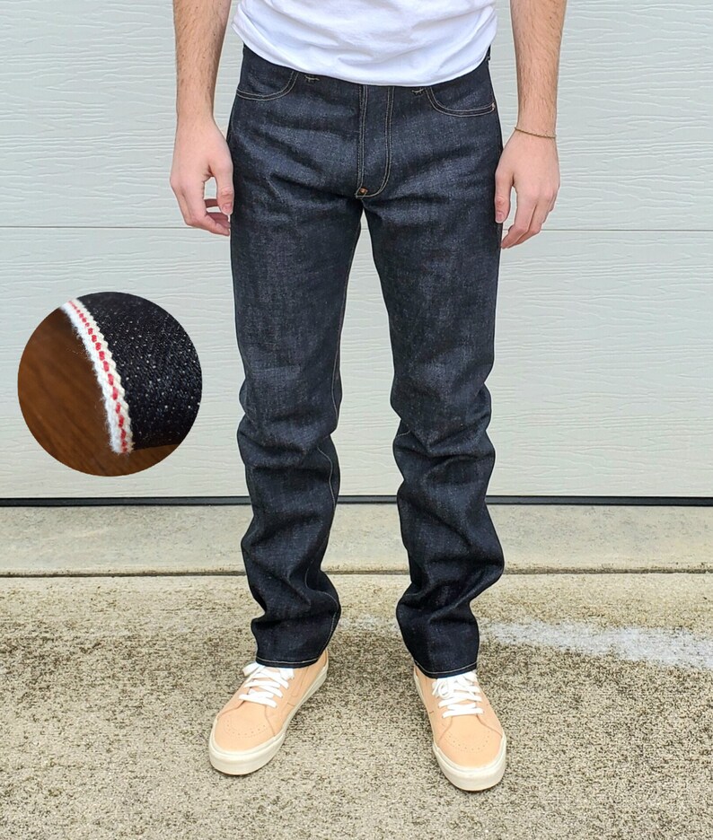 May include: Dark wash denim jeans with a classic five-pocket design. The jeans have a straight leg fit and are paired with tan sneakers. A close-up shows the selvedge detail with red and white stitching.