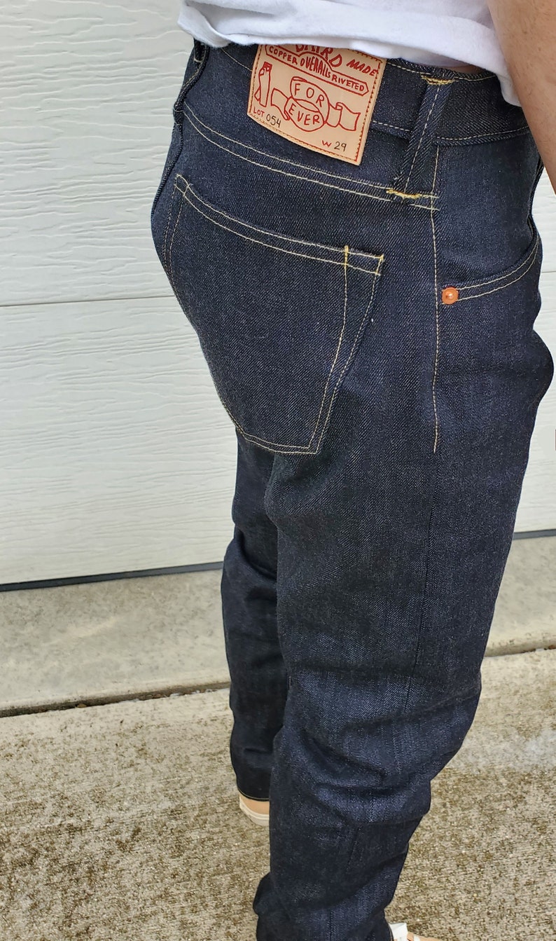 May include: Dark blue denim jeans with yellow stitching and a leather patch that reads "Copper Overalls Riveted For Ever." The jeans have a W29 size label and orange rivets.