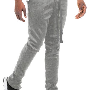 May include: Gray sweatpants with a drawstring waist and side ankle zippers. The pants are paired with black and white sneakers. The model is wearing a white t-shirt.