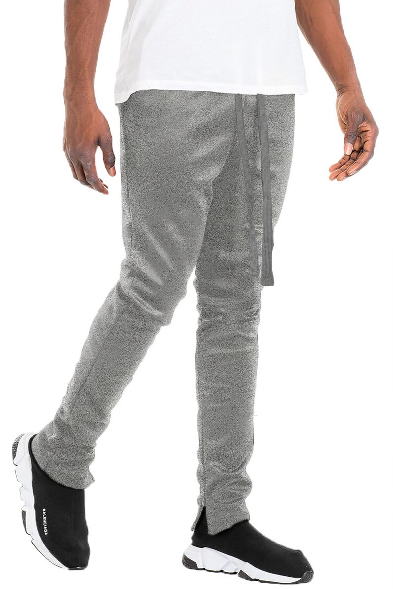 May include: Gray sweatpants with a drawstring waist and side ankle zippers. The pants are paired with black and white sneakers. The model is wearing a white t-shirt.
