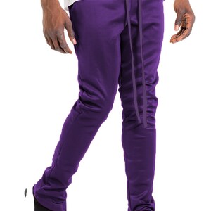 May include: Purple track pants with a drawstring waist and ankle zippers. The pants are a solid color and appear to be made of a smooth, possibly athletic material. The model is wearing black and white sneakers.
