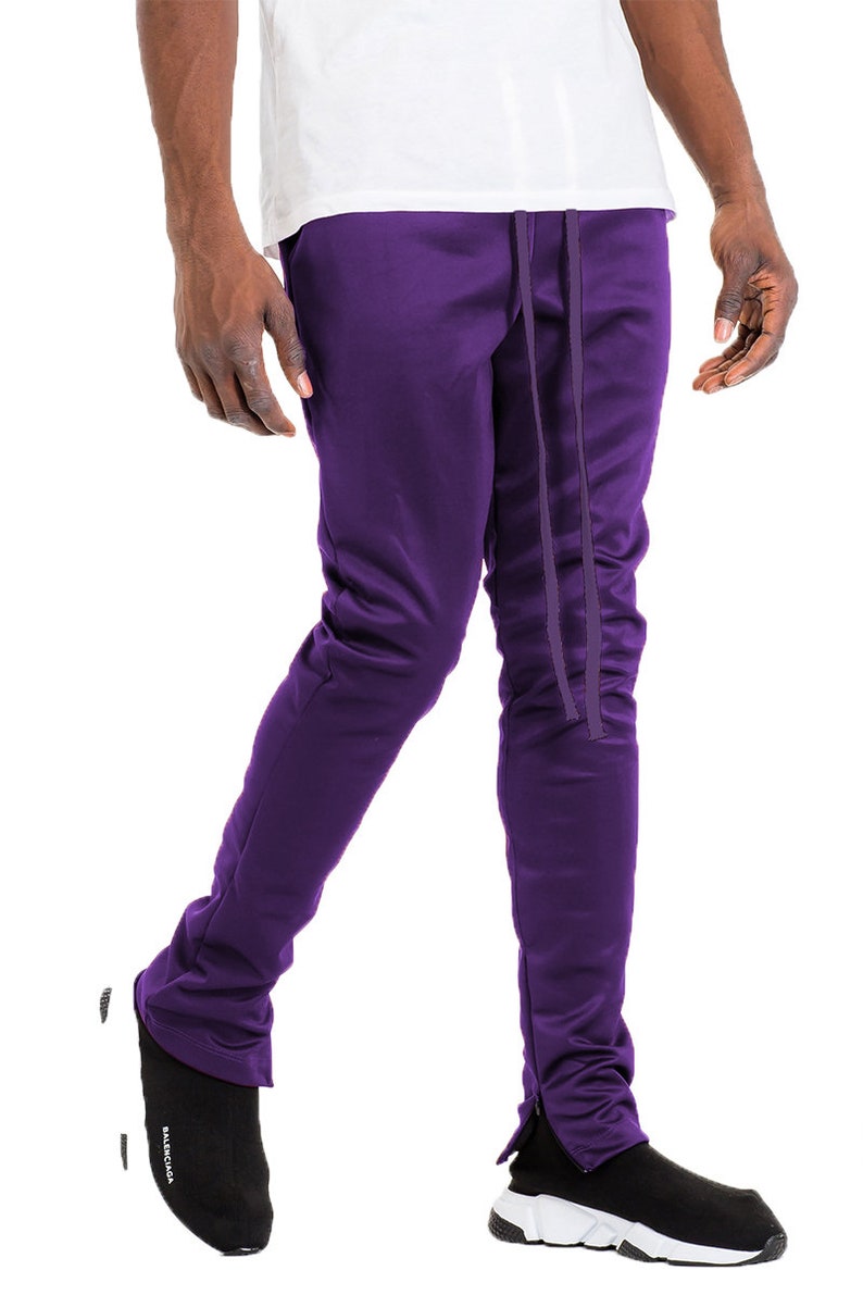 May include: Purple track pants with a drawstring waist and ankle zippers. The pants are a solid color and appear to be made of a smooth, possibly athletic material. The model is wearing black and white sneakers.