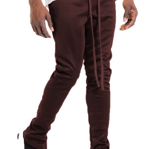 May include: Dark burgundy track pants with a drawstring waist and side zippers. The pants are paired with black and white athletic shoes. The pants are made of a smooth, possibly synthetic material.