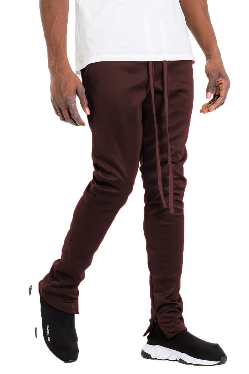 May include: Dark burgundy track pants with a drawstring waist and side zippers. The pants are paired with black and white athletic shoes. The pants are made of a smooth, possibly synthetic material.
