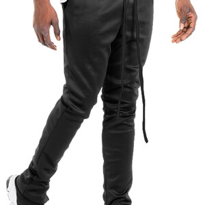 May include: Black satin pants with a drawstring waist and ankle zippers. The pants are paired with black and white sneakers. The pants have a slim fit and a sleek appearance.