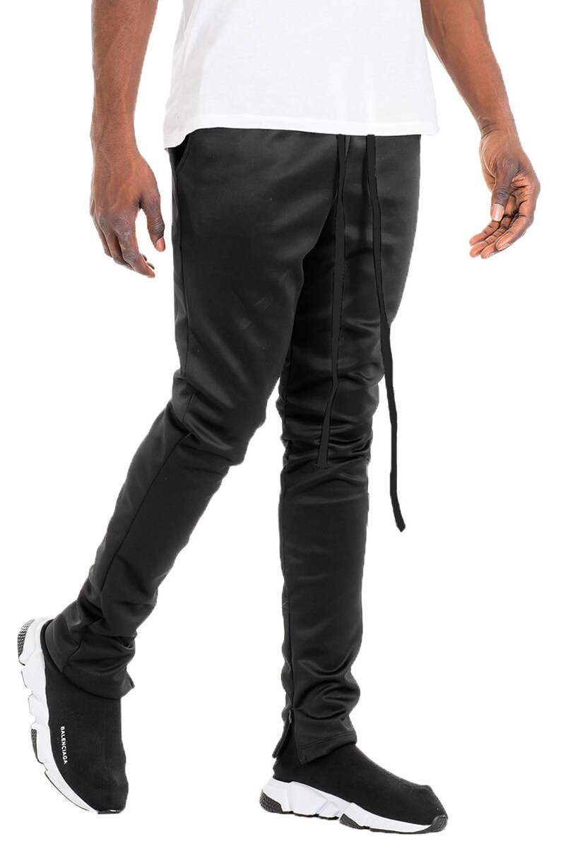 May include: Black satin pants with a drawstring waist and ankle zippers. The pants are paired with black and white sneakers. The pants have a slim fit and a sleek appearance.