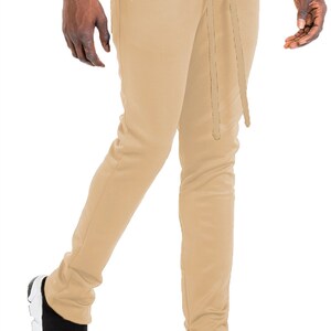 May include: Tan-colored sweatpants with a drawstring waist and ankle zippers. The pants are paired with black and white sneakers. The model is wearing a white t-shirt.