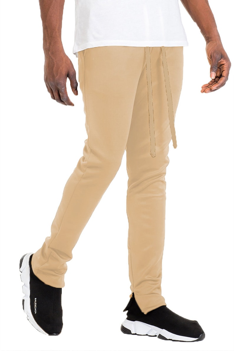 May include: Tan-colored sweatpants with a drawstring waist and ankle zippers. The pants are paired with black and white sneakers. The model is wearing a white t-shirt.