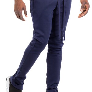 May include: Navy blue sweatpants with a drawstring waist and ankle zippers. The pants are paired with black and white sneakers. The model is wearing a white t-shirt.