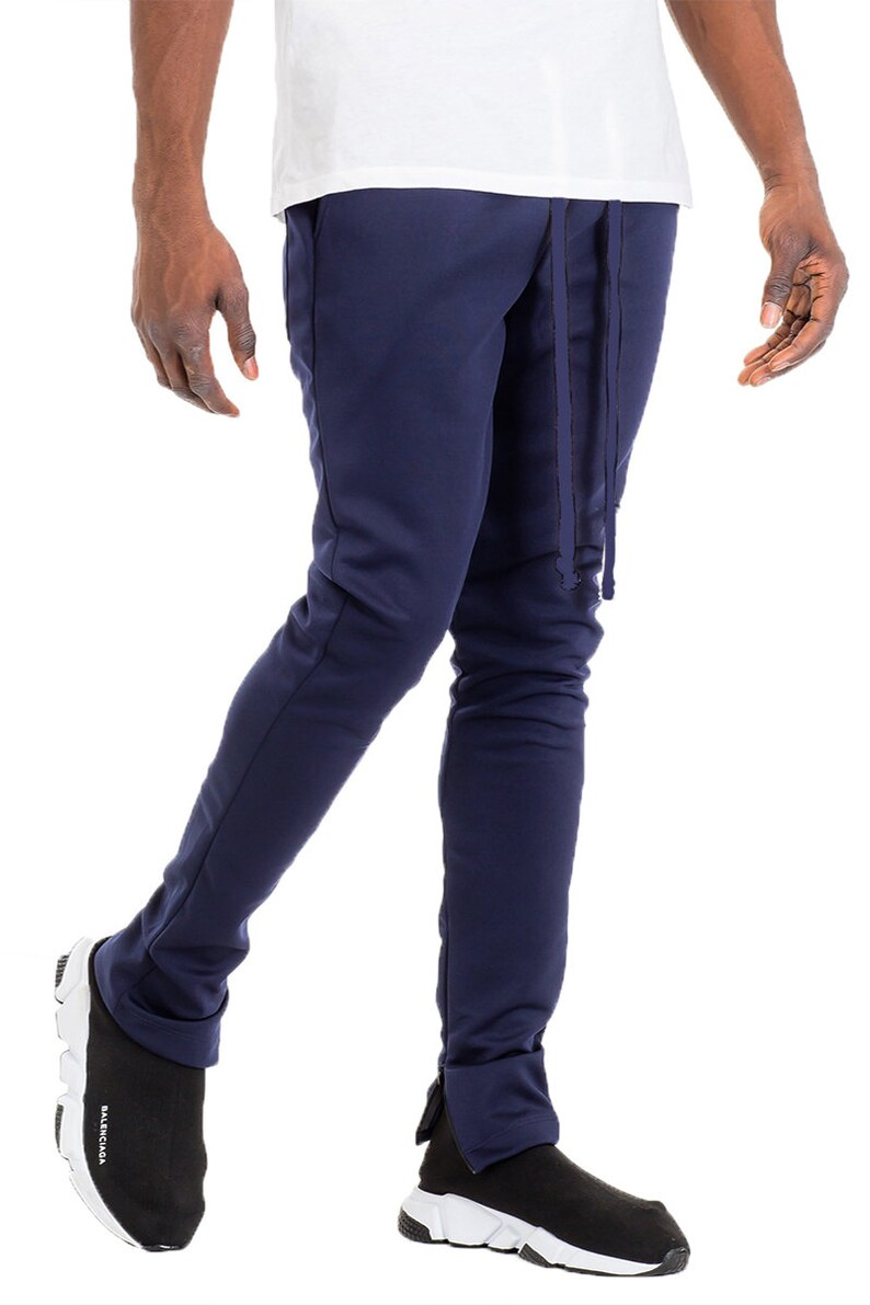 May include: Navy blue sweatpants with a drawstring waist and ankle zippers. The pants are paired with black and white sneakers. The model is wearing a white t-shirt.