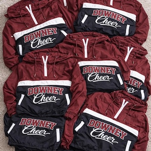 May include: A collection of maroon and black windbreakers with white accents. Each jacket features a hood, a half-zip closure, and the text "DOWNY Cheer" in white script across the chest. The jackets are arranged on a neutral-colored surface.