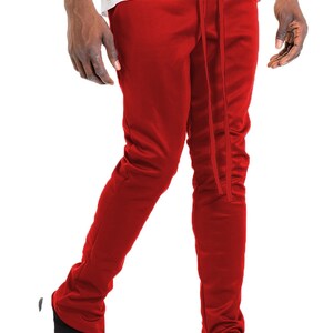 May include: Red track pants with a drawstring waist and ankle zippers. The pants are a solid color and appear to be made of a smooth, possibly athletic material. The pants are paired with black sneakers.