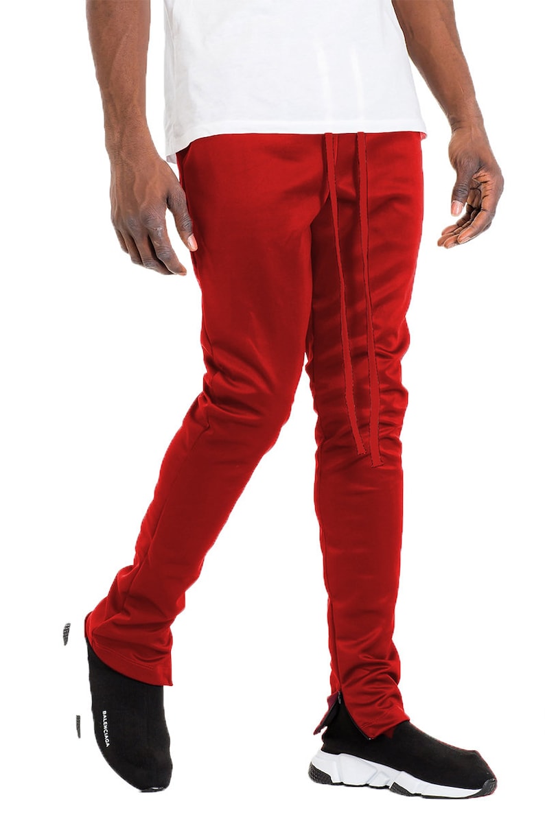May include: Red track pants with a drawstring waist and ankle zippers. The pants are a solid color and appear to be made of a smooth, possibly athletic material. The pants are paired with black sneakers.
