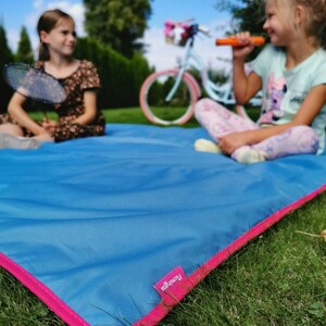 May include: Two girls are sitting on a blue picnic blanket with pink trim. The blanket has the word "Francoo" printed on it. One girl is holding a badminton racket and the other is holding a toy.