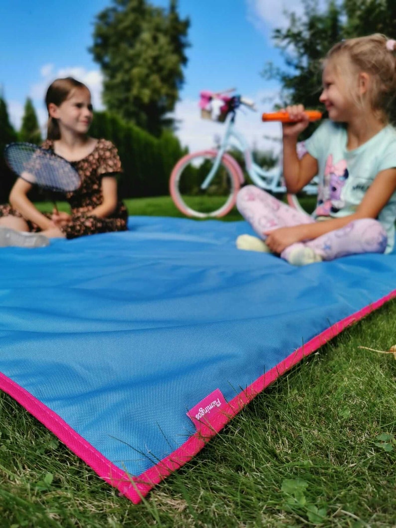 May include: Two girls are sitting on a blue picnic blanket with pink trim. The blanket has the word "Francoo" printed on it. One girl is holding a badminton racket and the other is holding a toy.