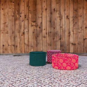 Könnte beinhalten: Drei runde Hocker mit verschiedenen gemusterten Stoffbezügen. Der Hocker links ist tief blaugrün, der mittlere Hocker hat ein geometrisches Muster in Rosa und Weiß, und der Hocker rechts hat ein rosafarbenes Blumenmuster mit gelben Akzenten.
