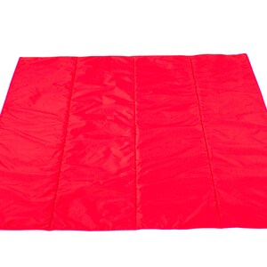 May include: A bright red quilted picnic blanket. The blanket is folded in half and has a pink label on the bottom left corner.