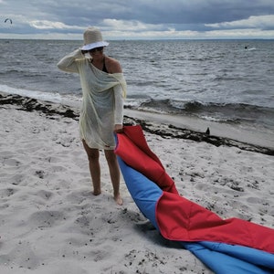 May include: A woman wearing a white hat and a yellow sheer cover-up is standing on a sandy beach. She is holding a red and blue beach blanket.