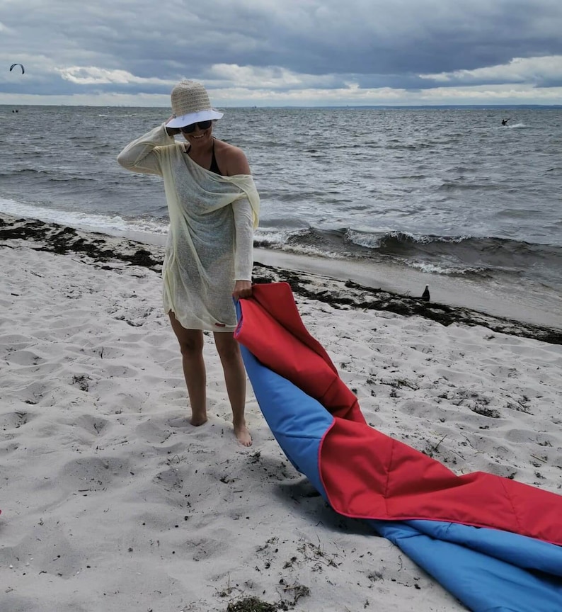 May include: A woman wearing a white hat and a yellow sheer cover-up is standing on a sandy beach. She is holding a red and blue beach blanket.