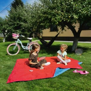 May include: Two children sit on a red and blue picnic blanket in a grassy yard. They are playing with a badminton racket and shuttlecock. A bicycle with a basket is parked in the background.