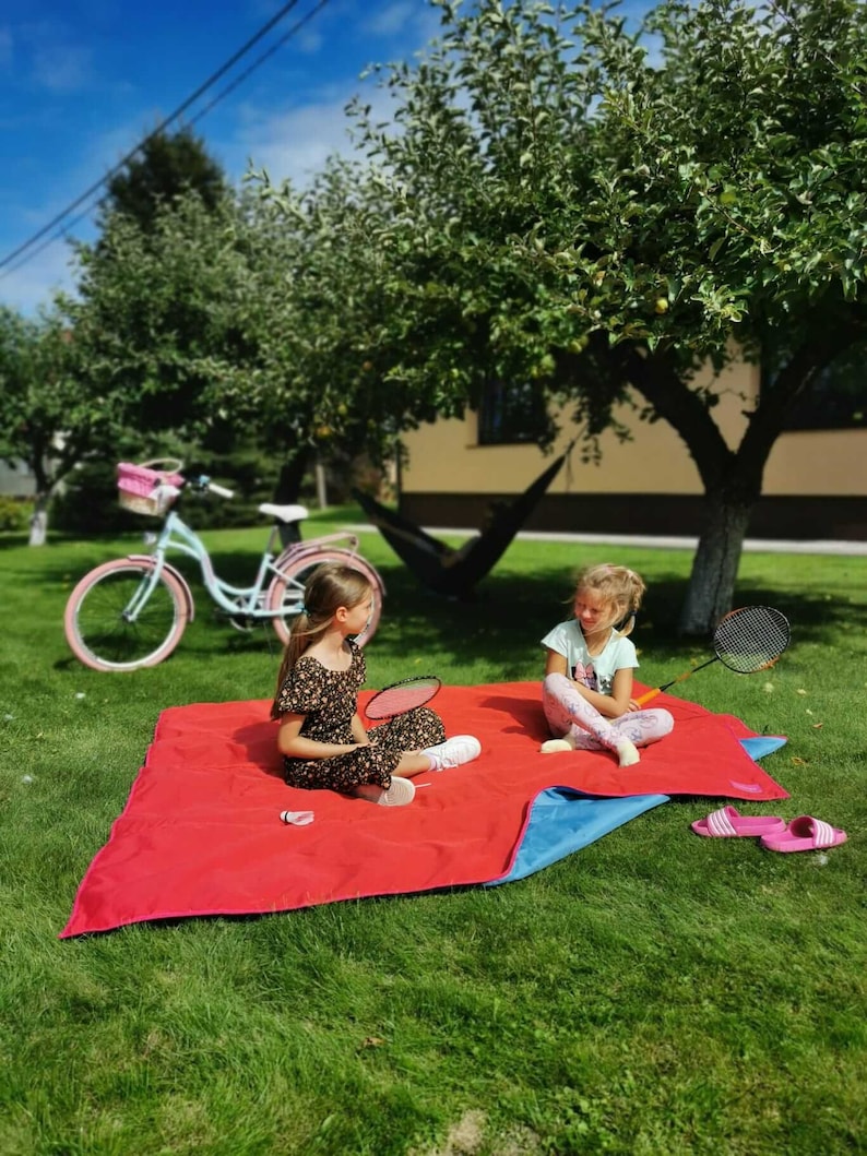 May include: Two children sit on a red and blue picnic blanket in a grassy yard. They are playing with a badminton racket and shuttlecock. A bicycle with a basket is parked in the background.