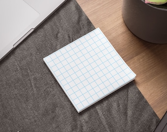 May include: A white gridded notepad with blue lines sits on a dark gray surface. A laptop and a potted plant are visible in the background.