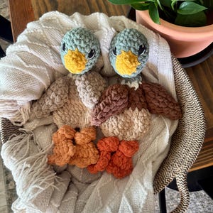May include: Handmade crochet ducklings with blue-green heads, yellow beaks, and black eyes. The ducklings are surrounded by crochet pieces in shades of brown, beige, and orange, all resting on a white cable-knit blanket.