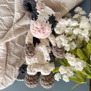 May include: A handmade crochet cow plushie with a pink muzzle, black and white patches, and brown hooves. The stuffed animal is lying on a cream-colored textured blanket, with white flowers and green stems in the background.
