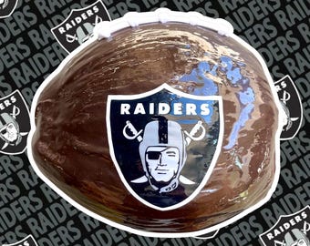 Puede incluir: Un pastel con forma de balón de fútbol, con una superficie marrón brillante y glaseado blanco en el borde superior. El pastel presenta el logotipo de los Raiders de Las Vegas en la parte delantera, un escudo con el nombre del equipo y una cara con casco.