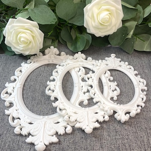 May include: Three white, oval picture frames with elaborate scrollwork and floral designs. They are arranged on a grey surface, with two white roses and green foliage in the background, creating a decorative display.