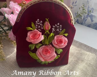 Könnte beinhalten: Eine bordeauxrote Samt-Münzbörse mit goldenem Rahmen und einem floralen Design, das mit rosa Satinband bestickt ist. Das Design zeigt drei rosa Rosen und grüne Blätter. Die Börse hat einen goldenen Verschluss und der Text "Amany Ribbon Arts" ist auf der Unterseite aufgedruckt.