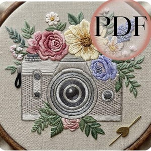May include: Embroidered design featuring a vintage camera surrounded by colorful flowers and green leaves. The camera is gray, with a black lens and strap. The flowers include pink roses, yellow daisies, and blue blossoms. The text "PDF" is in a pink circle.