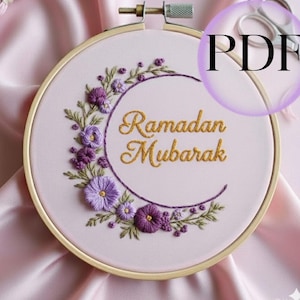 May include: A light pink embroidery hoop with the words "Ramadan Mubarak" in gold thread. The design features a crescent moon shape with purple flowers and green leaves. The hoop is made of wood.