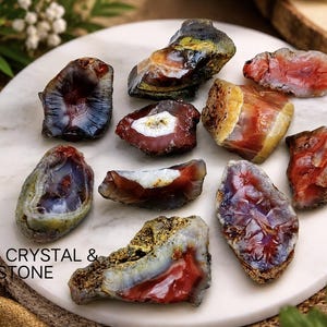 May include: A collection of ten raw, polished agate stone slices featuring vibrant red, orange, yellow, and deep purple hues with translucent centres. The stones are arranged on a white marble surface with the text SAMIR CRYSTAL & STONE printed.