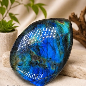 May include: A polished labradorite stone with a vibrant blue and green iridescent sheen, featuring natural dark veining and a smooth, rounded shape. The stone is labelled with the dimensions 70x50 mm and a weight of 150 grams.