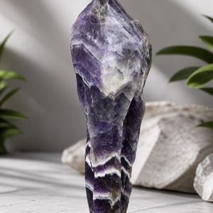 Könnte beinhalten: Ein großer, roher Amethystkristall mit einem geschichteten, gebänderten Aussehen. Der Kristall weist Farbtöne von tiefem Lila, Lavendel und Weiß auf. Der Naturstein steht aufrecht vor einem neutralen Hintergrund und zeigt seine einzigartige geologische Formation.