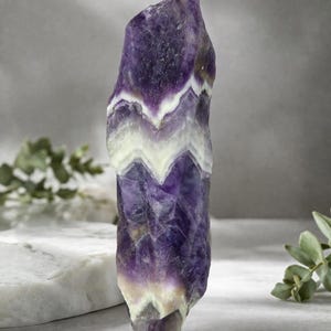 Könnte beinhalten: Ein roher Amethystkristall mit einem geschichteten Chevron-Muster in tiefem Lila, Weiß und blassem Gelb. Der natürliche Edelstein steht aufrecht und zeigt seine einzigartigen Farbvariationen und seine kristalline Struktur. Der Kristall ist ein dekorativer Gegenstand.