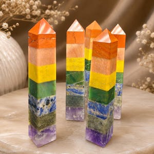 Può includere: Una collezione di cinque torri di cristallo verticali, ciascuna composta da sette segmenti di pietra impilati di colori distinti tra cui arancione, giallo, verde, blu e viola, disposti in una forma a prisma esagonale appuntita con finitura lucida.
