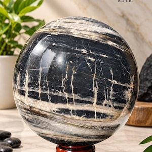 Puede incluir: Una piedra esférica pulida con patrones veteados en blanco y negro, apoyada sobre un pequeño soporte circular de madera. El texto en la imagen indica un tamaño de 210 mm, un peso de 12.150 gramos y 8,5 pulgadas, reflejando sus dimensiones.