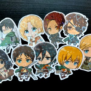 May include: A collection of nine die-cut stickers featuring chibi-style anime characters from Attack on Titan. Each sticker displays a unique character with large eyes and distinct hair colors, including blonde, brown, and black, with a holographic finish.