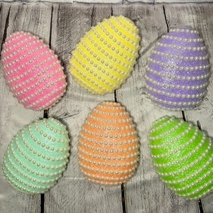 May include: Six decorative Easter eggs in various colors: pink, yellow, purple, mint green, orange, and lime green. Each egg is covered in glitter and adorned with rows of small, white pearls. The eggs are arranged on a weathered, light-colored wooden surface.