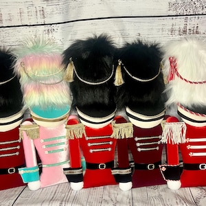 May include: Five plush nutcracker figures in a row, each wearing a tall fuzzy hat with a chin strap. The figures feature velvet-like jackets in red, pink, and dark red, adorned with gold or white fringe epaulets, horizontal trim, and black belts.