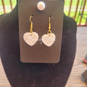 May include: White heart-shaped earrings with gold-colored hooks. The earrings are displayed on a black card with a pearl stud earring at the top. The text "BeadsbyStorm" is written on the earrings.
