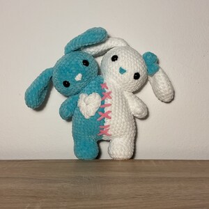 May include: Two crocheted bunny plushies, one blue and one white, are sewn together to create a single plush toy. The blue bunny has a pink heart on its chest, and the white bunny has a pink stitched line down its back.