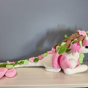 May include: A handmade white crochet dragon plush toy with pink and green floral accents. The dragon features a long tail, large pink wings, and a brown horn-like structure on its head. The dragon is lying on a light-colored surface.